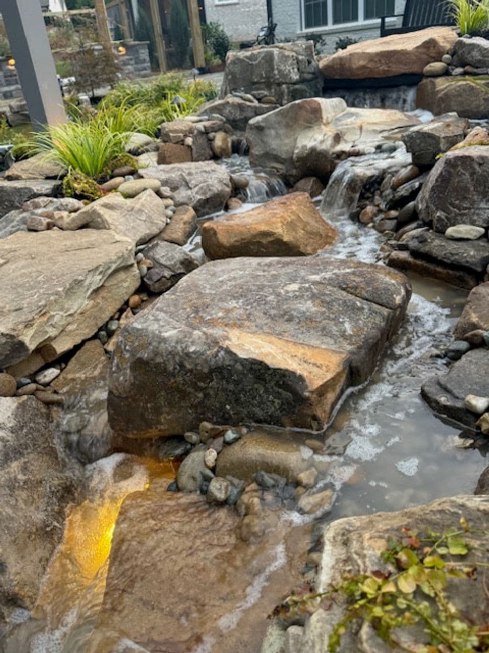 Natural stone water feature with flowing water and lush greenery in a landscaped setting.