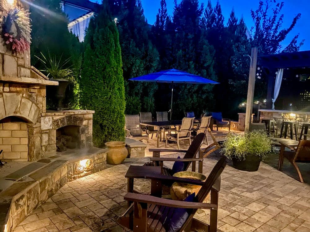 Cozy outdoor patio at dusk with seating, umbrella, and fireplace surrounded by greenery.