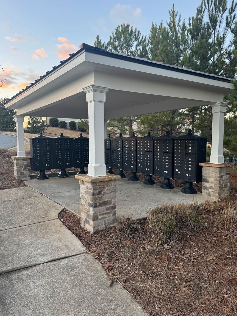 Black cluster mailboxes under a roof near a residential area, surrounded by greenery.