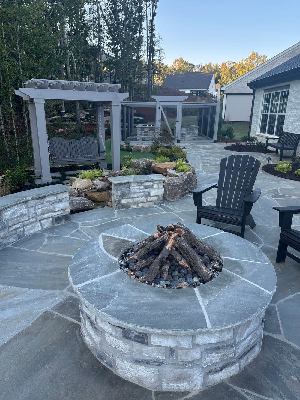 Modern stone fire pit with wooden chairs in landscaped backyard, surrounded by greenery.