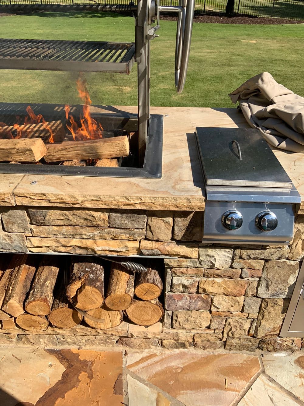 Outdoor stone grill featuring flames, firewood storage, and a stainless steel side burner.
